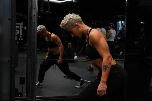 a lifter in a black sports bra and black joggers walking up to a loaded barbell on the floor of a dark gym rack, looking down at it