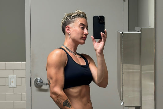lifter in a black sports bra photographed in a gym restroom mirror, shoulder and arm development visible in profile