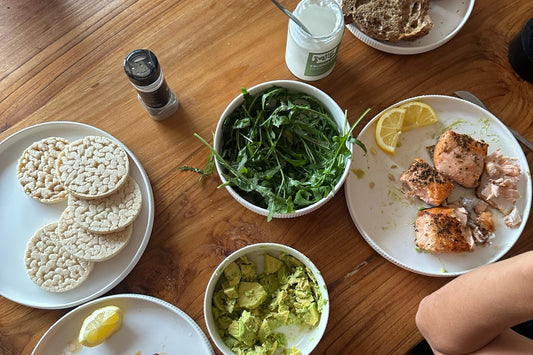 a wooden table seen from above with plates of salmon, rice cakes, a bowl of rocket, mashed avocado, coconut yoghurt and a lemon wedge