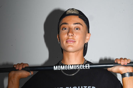 lifter in a backwards cap holding a black MoMo Muscle barbell across the shoulders in a grey studio