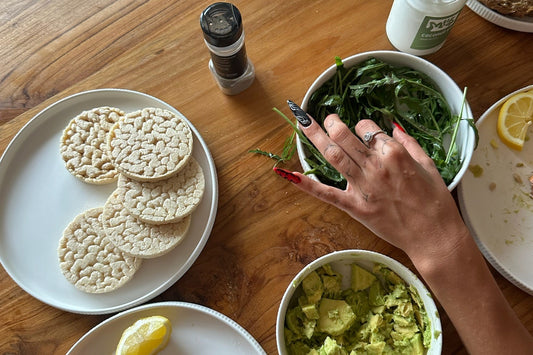 overhead view of a wooden table with plates of salmon, rice cakes, arugula, avocado and yogurt