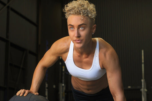 lifter in a white sports bra doing a push-up with one hand on a medicine ball in a dark gym, looking up