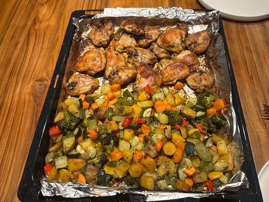 a roasting tray on a wooden table holding baked chicken thighs and a large pile of roasted broccoli, carrots, courgette, peppers, potatoes and onion