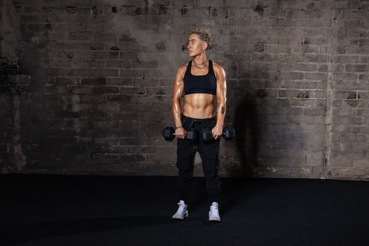 Morgan Olson standing against a dark brick wall holding a dumbbell in each hand
