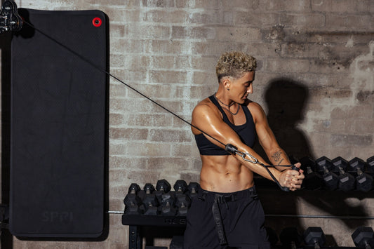 Morgan Olson pulling a loaded cable across her body in a dark brick studio, arms locked, face set