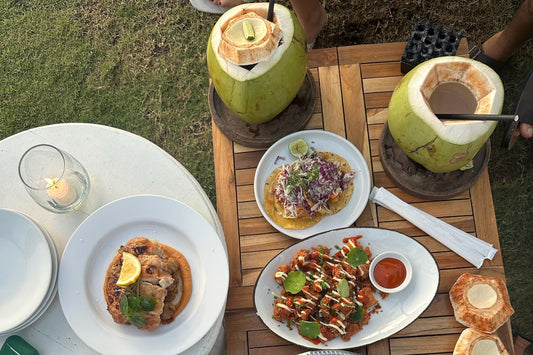 Overhead view of a wooden table outdoors with two fresh coconuts with straws, a plate of grilled chicken, tacos, a plate of fried seafood and small coconut bowls