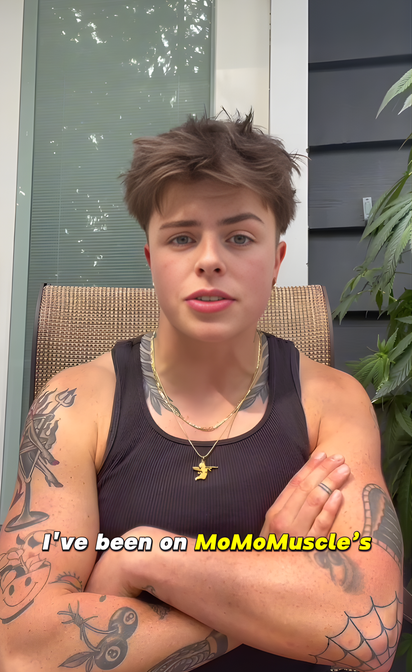 Person with tattoos wearing a black tank top, standing outdoors.